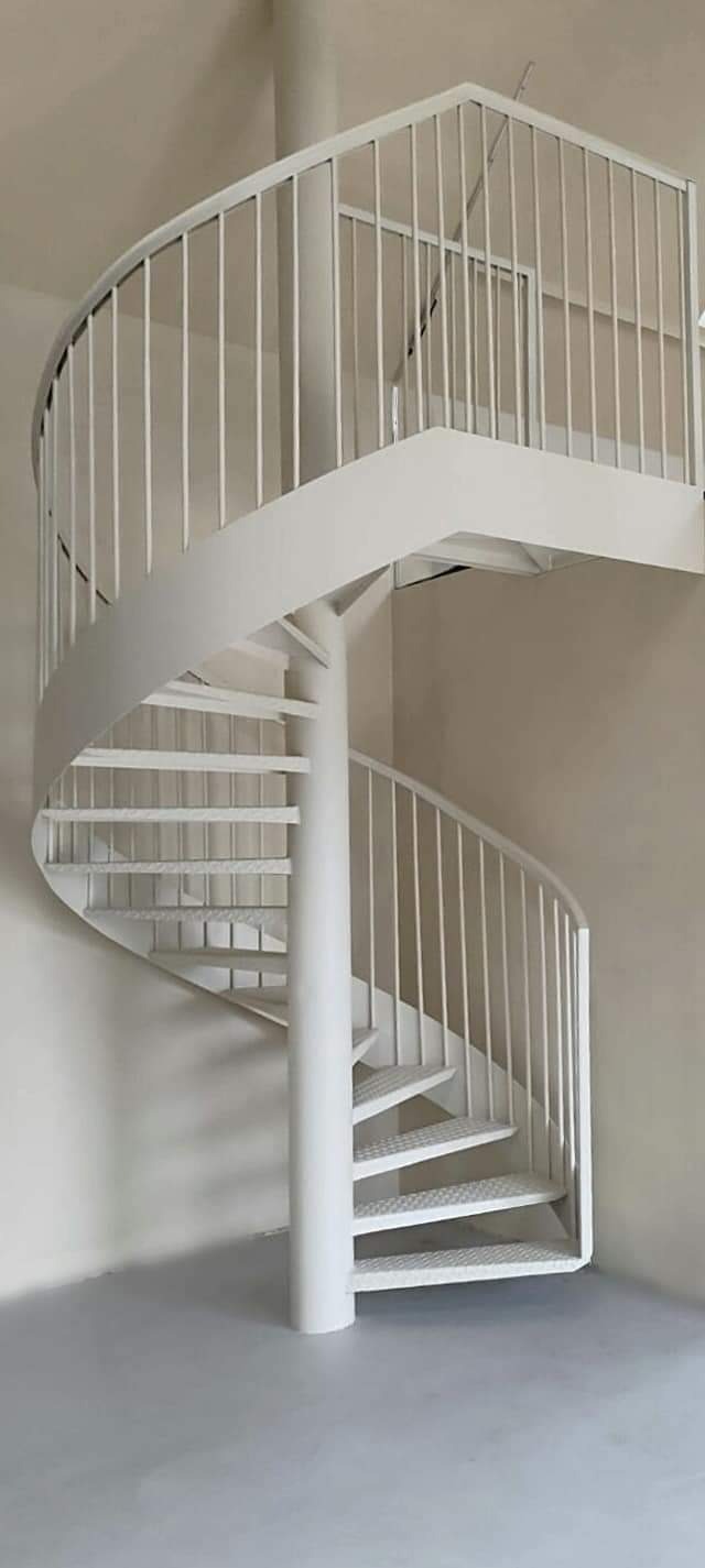 Białe metalowe schody spiralne z perforowanymi stopniami i poręczą z pionowych prętów, umieszczone w jasnym pomieszczeniu z kremowymi ścianami.