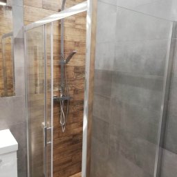 MAROBUD Marek Furtak - Nowoczesna kabina prysznicowa z przesuwnymi drzwiami, wyłożona drewnopodobnymi płytkami na ścianie prysznica i szarymi płytkami na pozostałych ścianach, widoczny zestaw prysznicowy z deszczownicą.