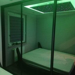 MAROBUD Marek Furtak - Sypialnia z zamontowanym sufitem podwieszanym z efektem gwiazd, widoczna przez przesuwne drzwi szafy w kolorze zielonym, z grzejnikiem pod oknem.