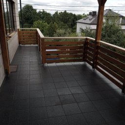MAROBUD Marek Furtak - Wykończony balkon z ciemną, kwadratową podłogą z gumowych płytek, drewnianą balustradą i częściowo zadaszeniem, widok na zieleń i dom w tle.