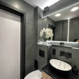 Nowoczesna łazienka w odcieniach szarości z białą armaturą, owalną umywalką nablatową, wiszącą toaletą i dekoracyjną białą orchideą na półce.