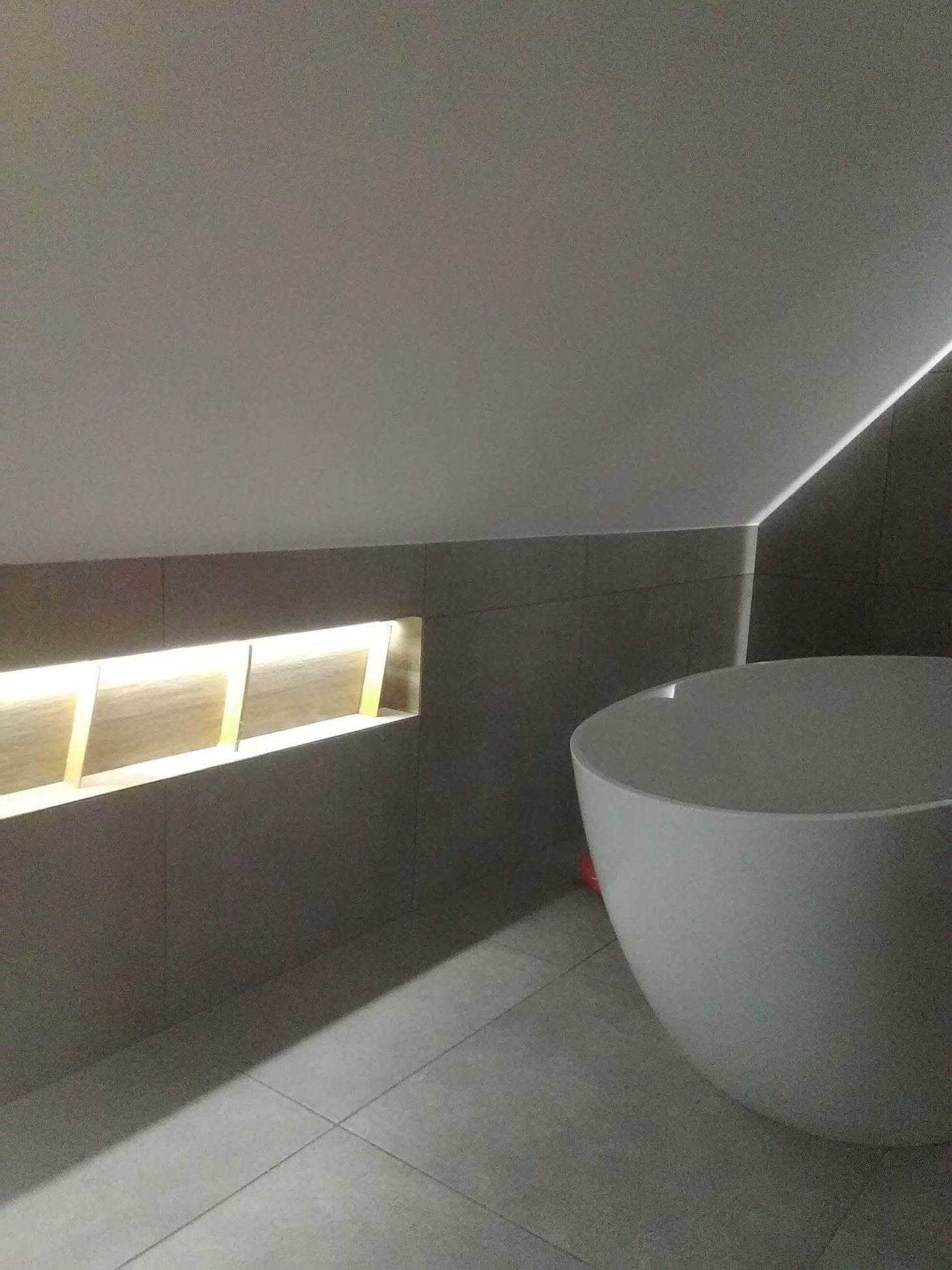Nowoczesna łazienka na poddaszu z owalną, białą wanną wolnostojącą, szarymi płytkami i podświetlaną wnęką w ścianie.