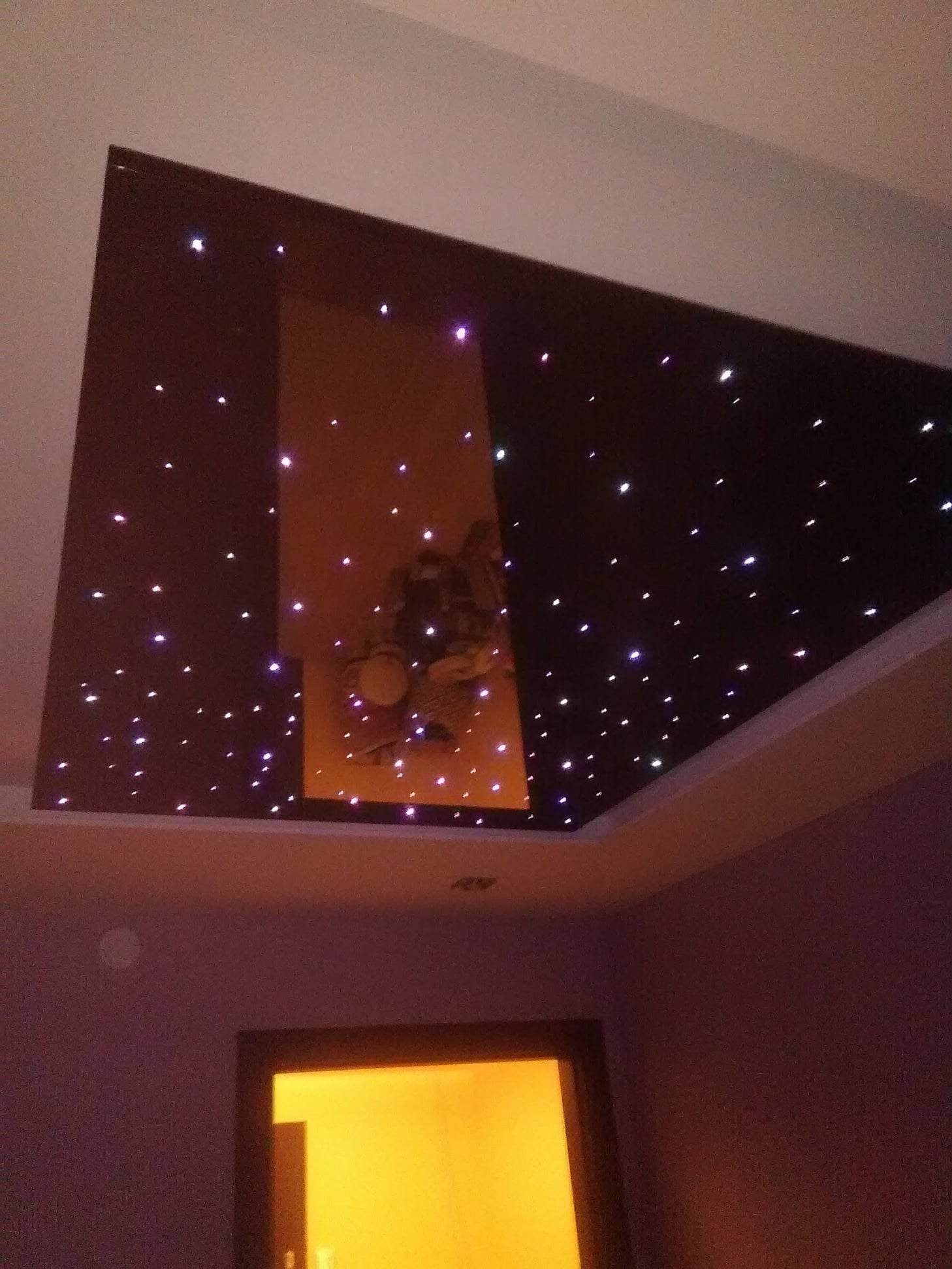Ciemnobrązowy sufit podwieszany z efektem rozgwieżdżonego nieba, gdzie białe punkty światła imitują gwiazdy, a w centrum znajduje się prostokątny panel z podświetlanym obrazem.