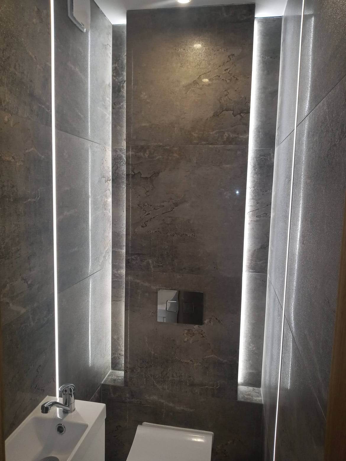 Nowoczesna łazienka z szarymi płytkami imitującymi kamień, podświetleniem LED w pionowych wnękach, minimalistyczną umywalką i podtynkową spłuczką WC.