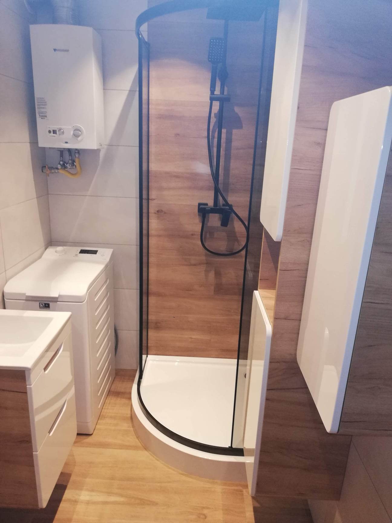 Nowoczesna łazienka z kabiną prysznicową z czarnymi profilami, umywalką z szafką w kolorze drewna, pralką i piecykiem gazowym na ścianie z białych płytek.