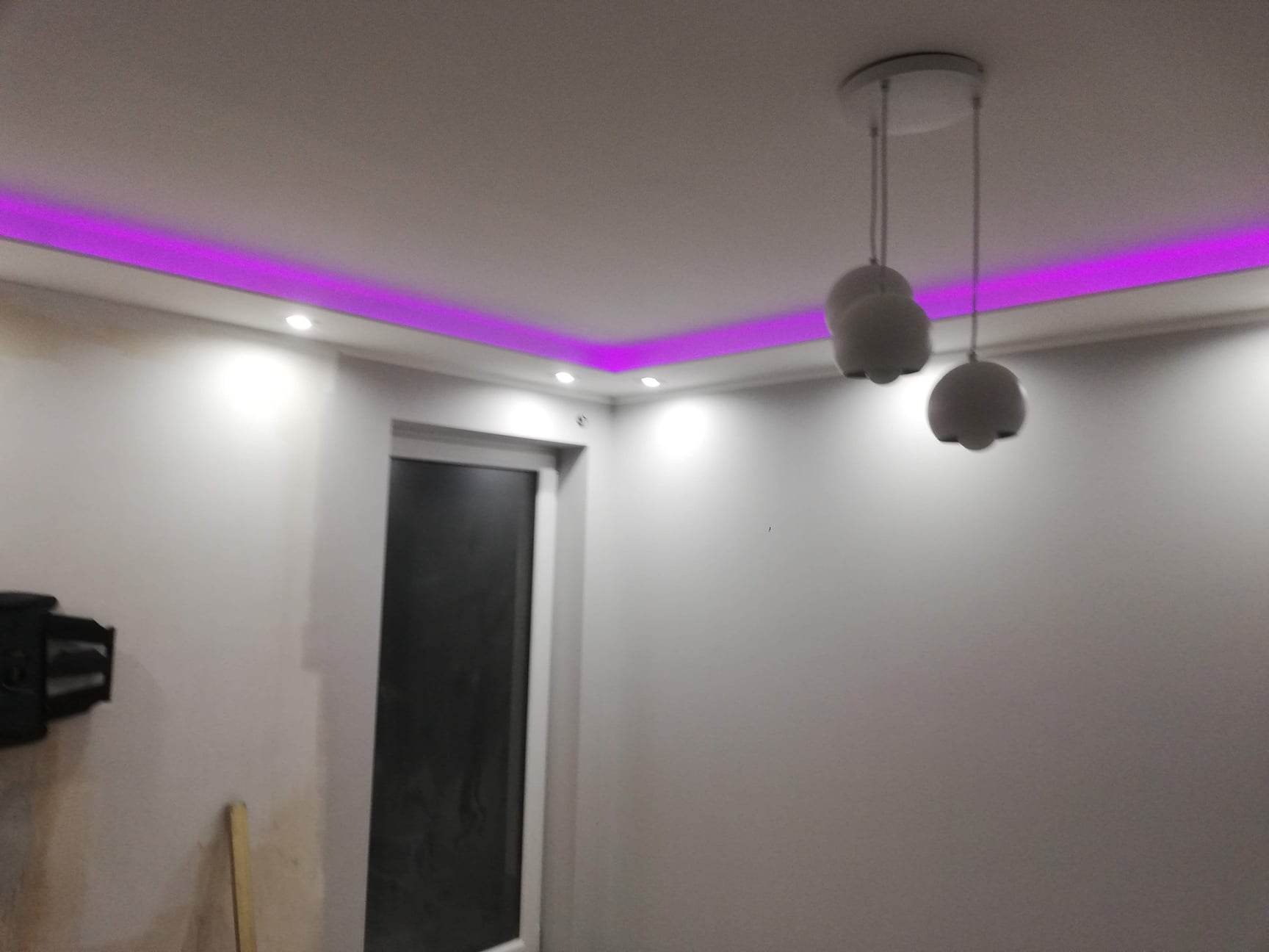 Pokój w trakcie wykańczania z pomalowanymi ścianami na biało, podwieszanym sufitem z fioletowym oświetleniem LED i nowoczesną lampą wiszącą; widoczne narzędzia malarskie w rogu.