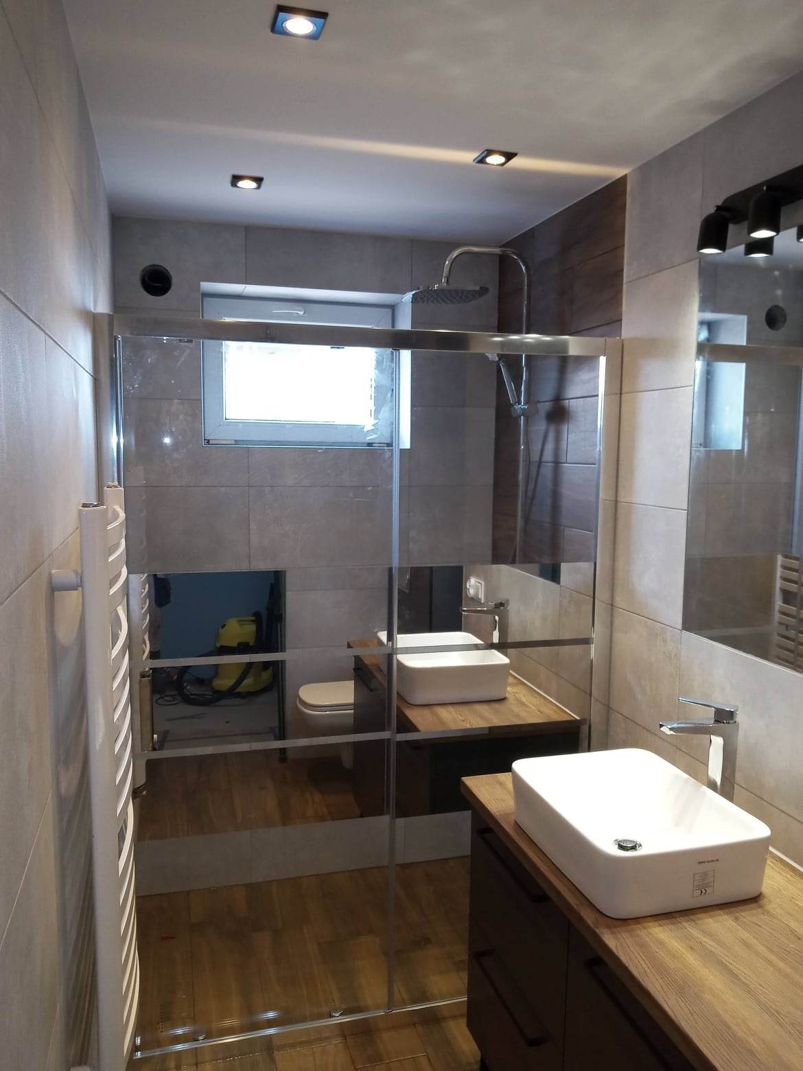 Wykończona łazienka z kabiną prysznicową ze szklaną ścianką, dwiema umywalkami nablatowymi na drewnianych blatach, grzejnikiem drabinkowym i widocznym przez szybę odkurzaczem przemysłowym.