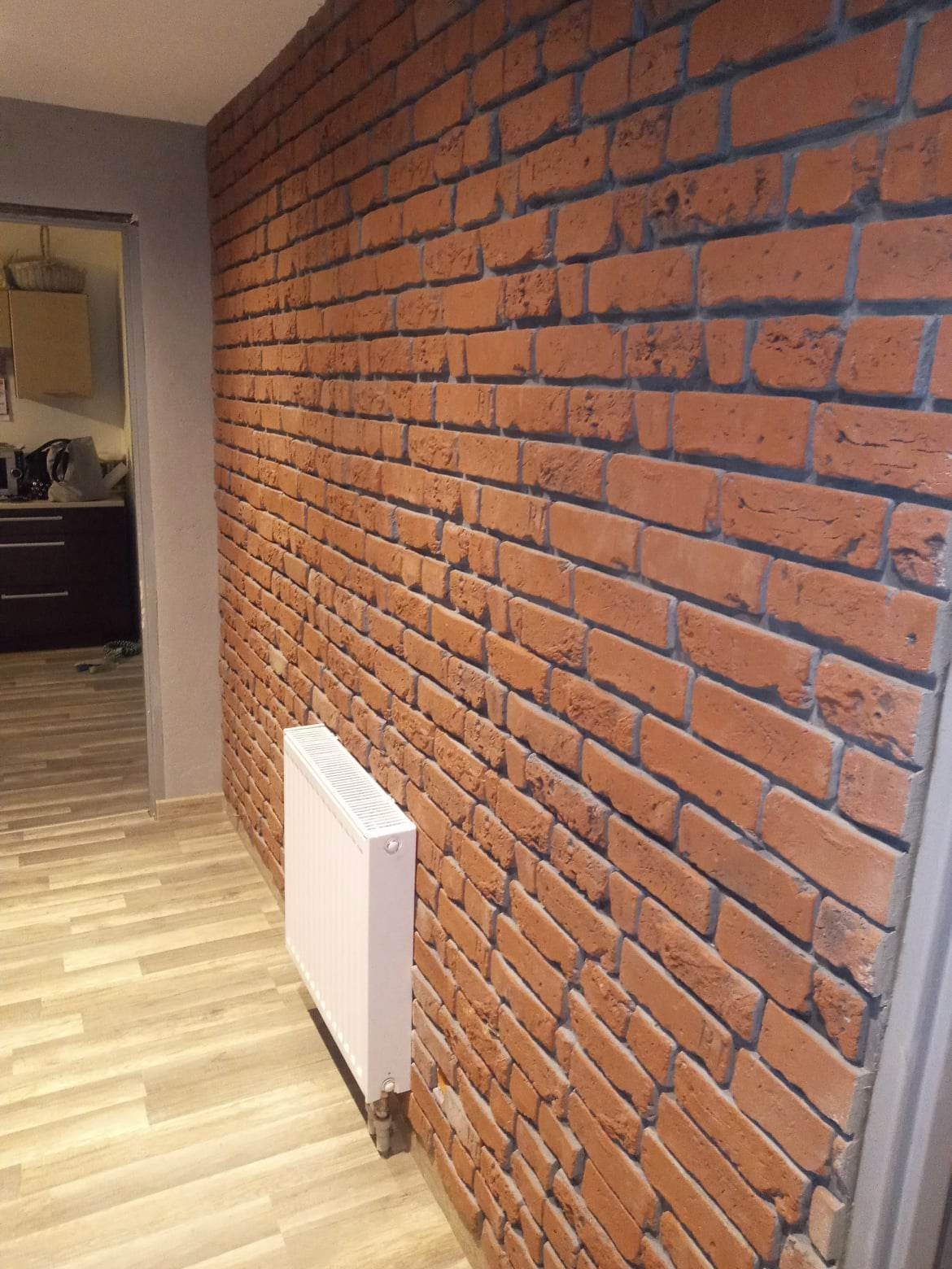 Ściana wyłożona dekoracyjną cegłą w odcieniach czerwieni i brązu z widocznym białym grzejnikiem, na podłodze panele imitujące drewno, widok z boku.