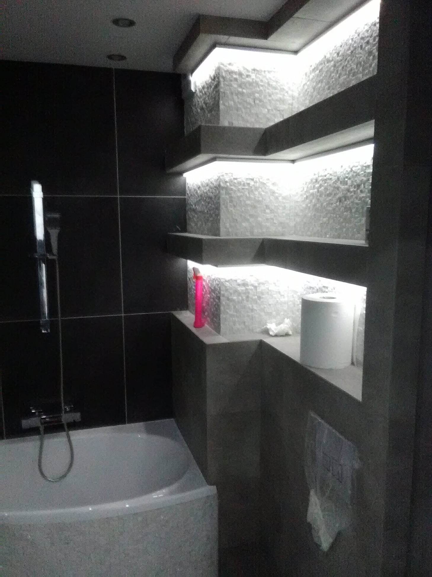 Nowoczesna łazienka z czarnymi płytkami i podświetlanymi półkami z mozaiką, widoczna wanna, armatura i akcesoria łazienkowe.