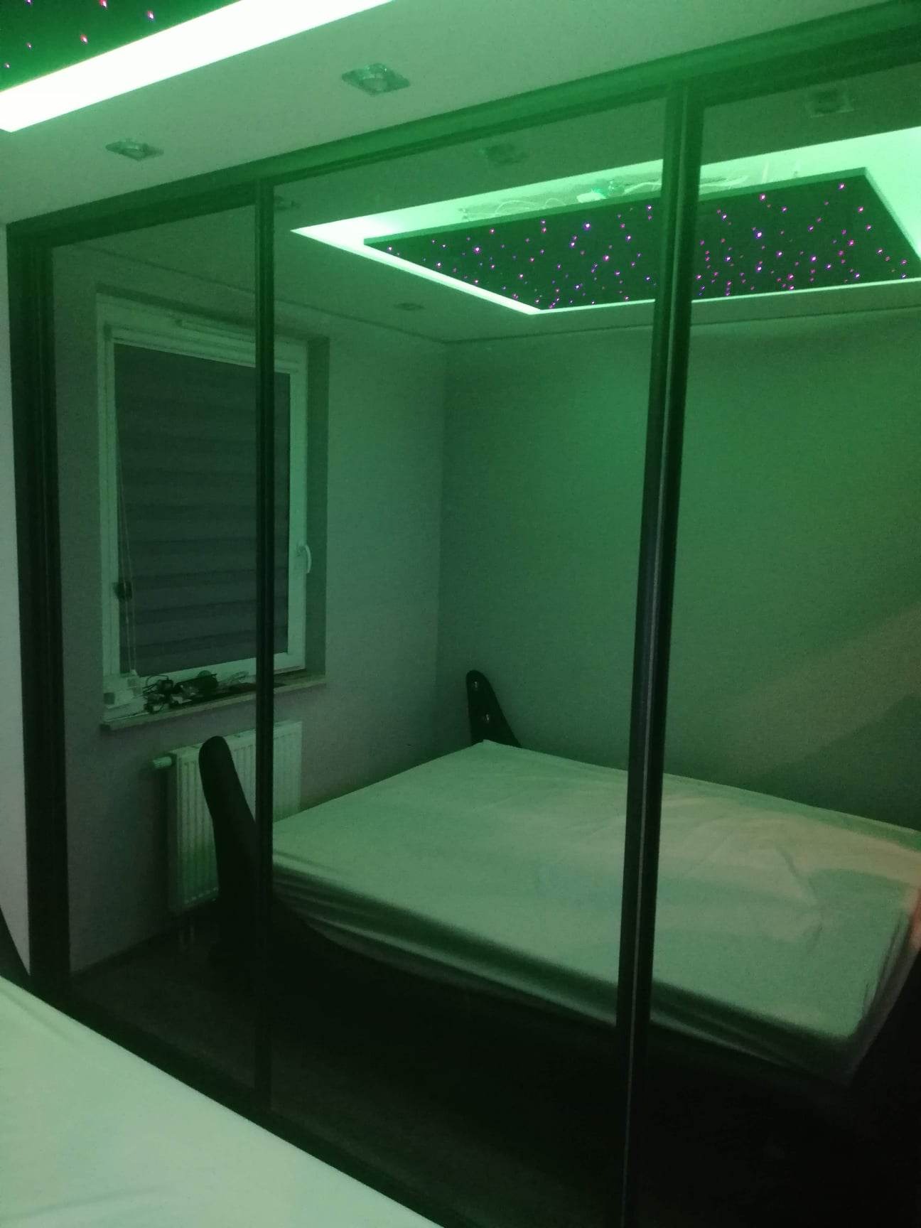 Sypialnia z zamontowanym sufitem podwieszanym z efektem gwiazd, widoczna przez przesuwne drzwi szafy w kolorze zielonym, z grzejnikiem pod oknem.