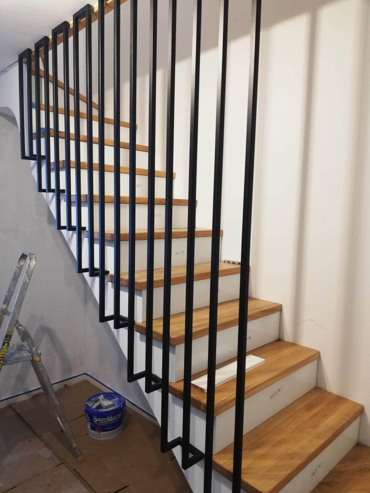 Schody drewniane z białymi podstopnicami i czarną, minimalistyczną balustradą, widoczne ślady prac malarskich, drabina i puszka farby Speedmaster w tle.