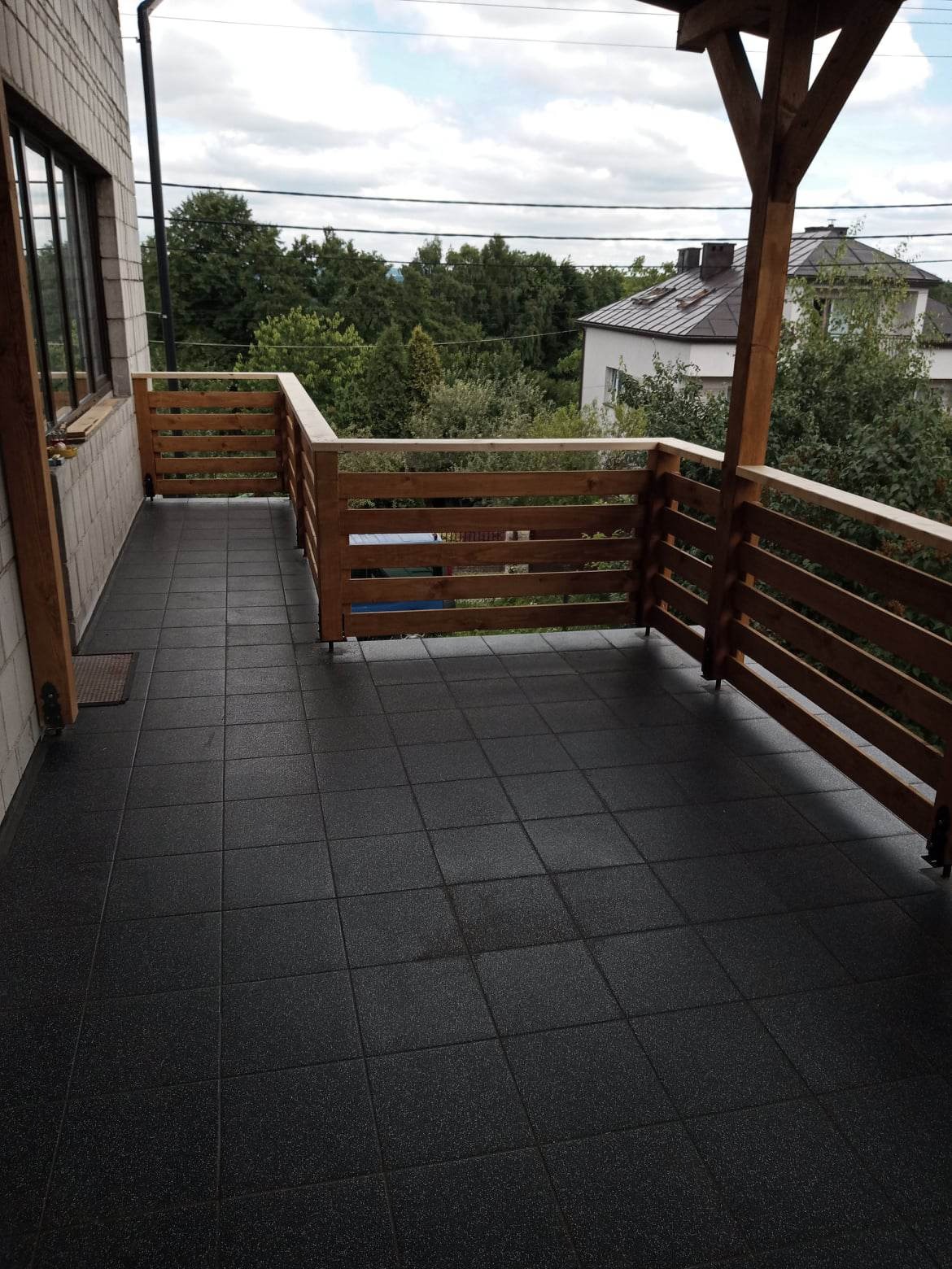 Wykończony balkon z ciemną, kwadratową podłogą z gumowych płytek, drewnianą balustradą i częściowo zadaszeniem, widok na zieleń i dom w tle.