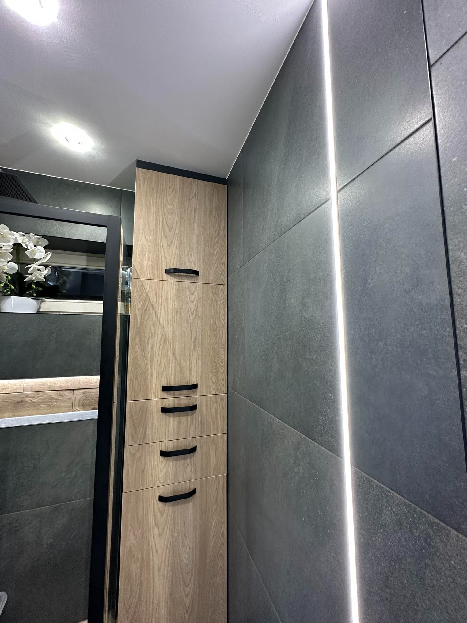 Nowoczesna łazienka z szarymi płytkami i pionowym oświetleniem LED, szafka z drewnopodobnym fornirem i czarnymi uchwytami, widoczne elementy dekoracyjne w lustrze.