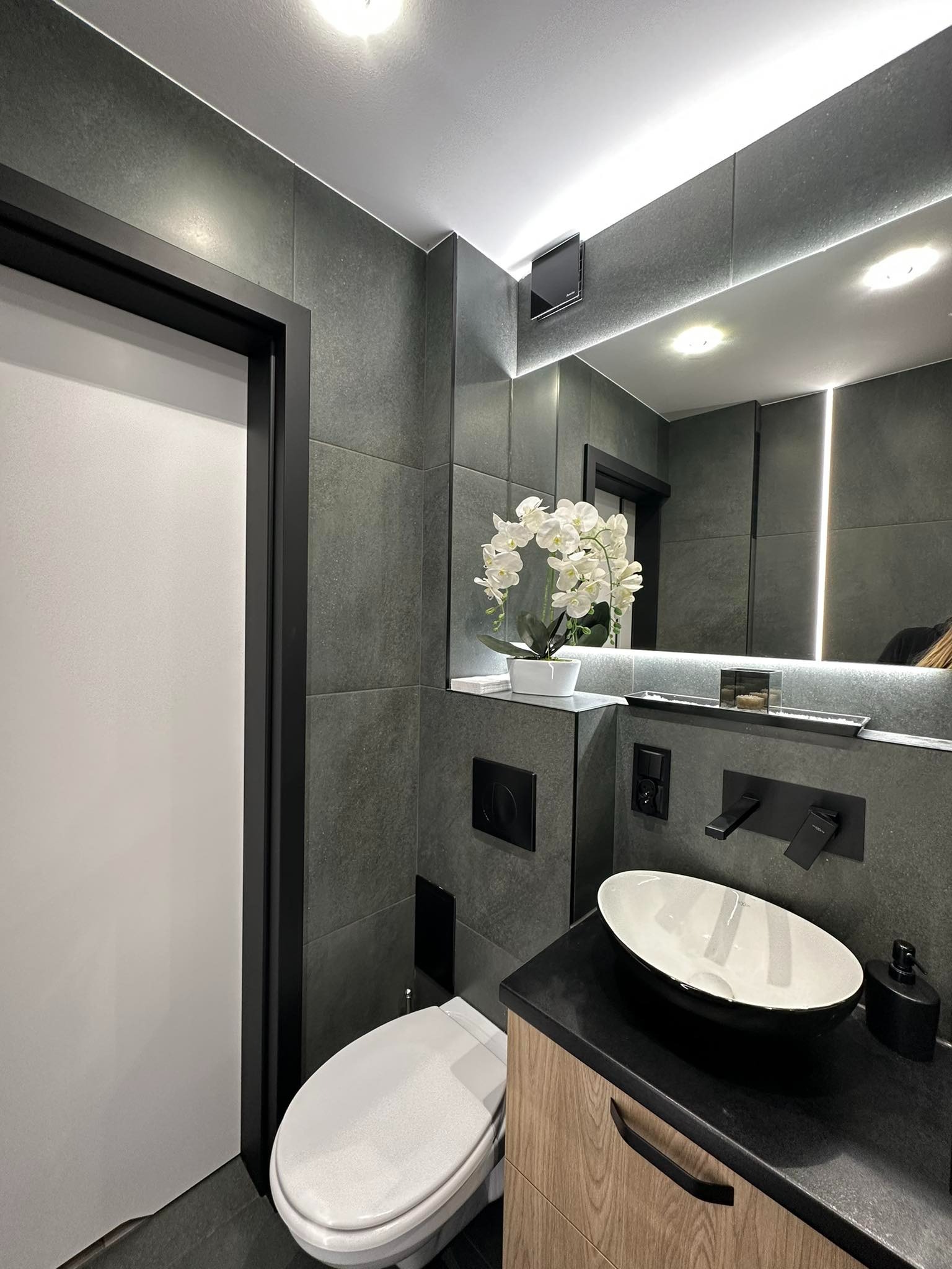 Nowoczesna łazienka w odcieniach szarości z białą armaturą, owalną umywalką nablatową, wiszącą toaletą i dekoracyjną białą orchideą na półce.