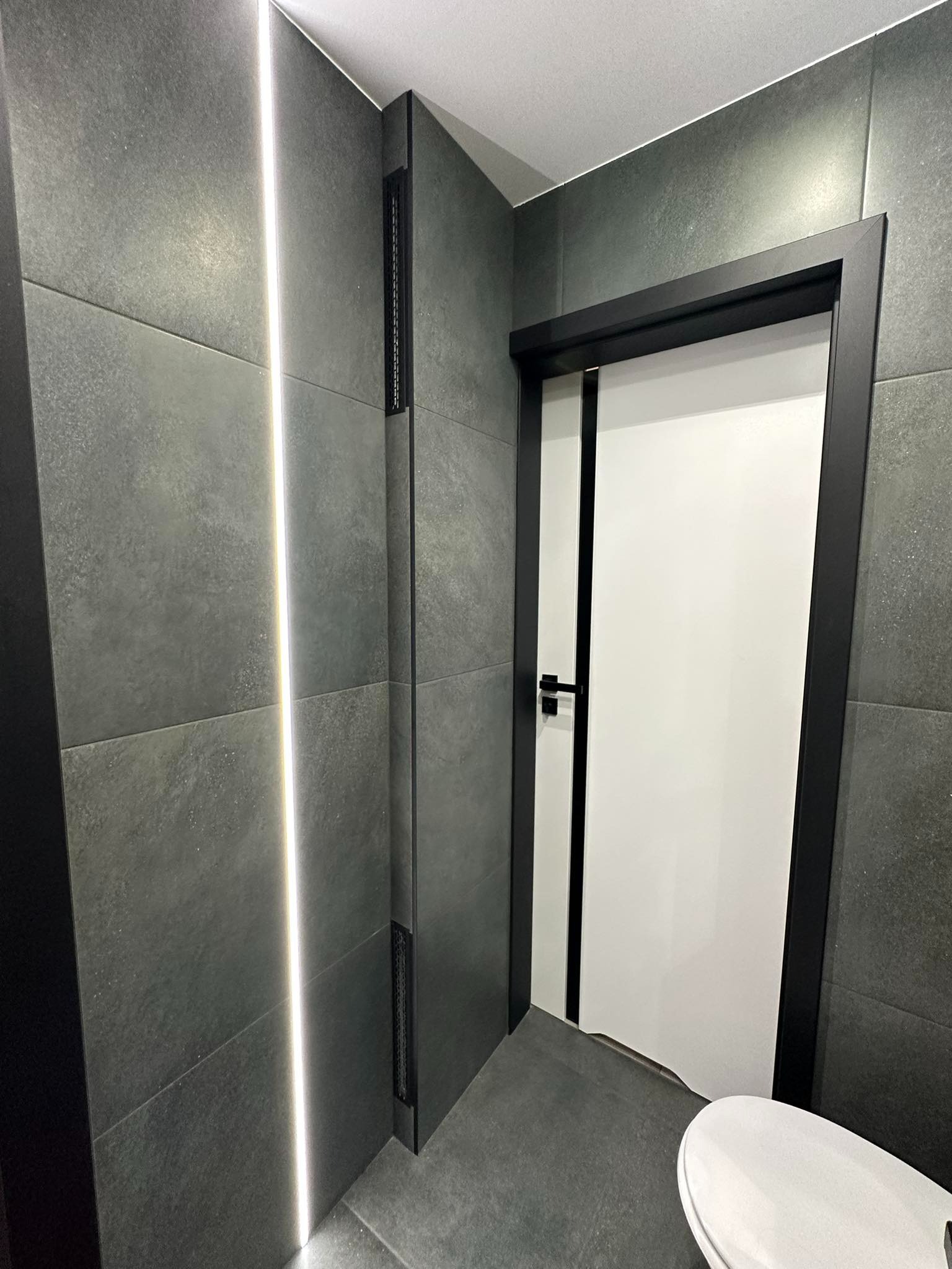 Nowoczesna łazienka z szarymi płytkami wielkoformatowymi, podświetleniem LED wzdłuż ściany i białą, minimalistyczną toaletą oraz drzwiami z czarną ramą.