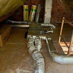 COLD FACTORY - Instalacja rekuperacji na poddaszu: srebrne, izolowane rury wentylacyjne prowadzą do centrali rekuperacyjnej, widoczne materiały izolacyjne Isover, ceglany komin i kartonowe pudło Chojniak.