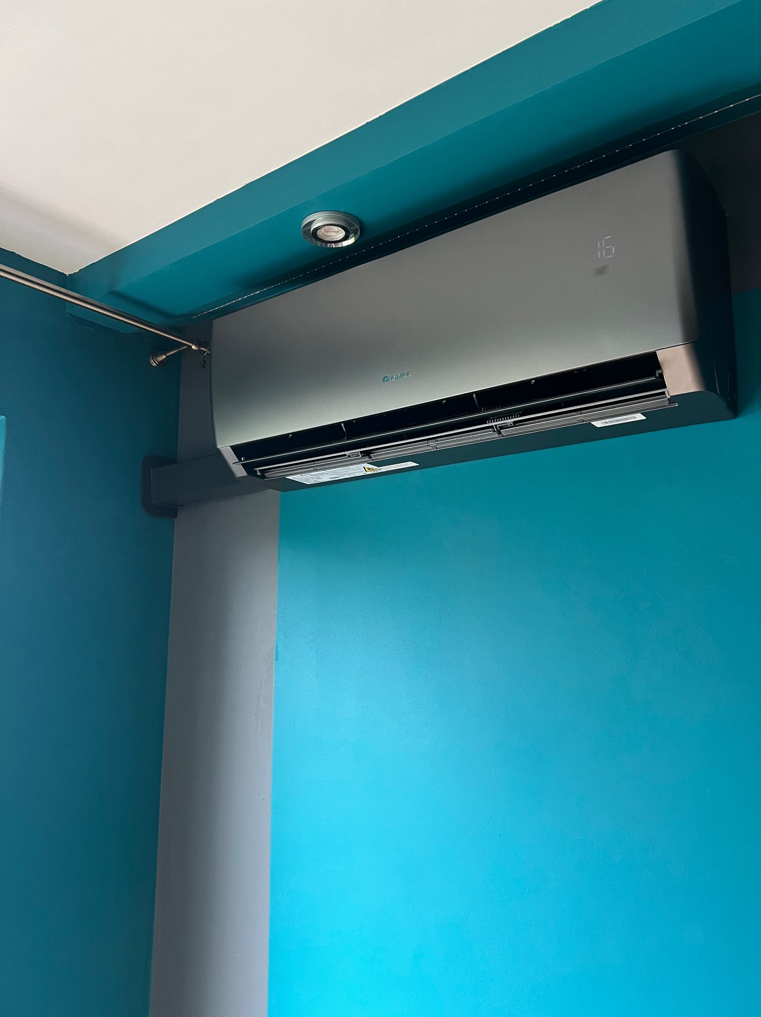 Nowoczesny, grafitowy klimatyzator marki Gree zamontowany na turkusowej ścianie w pomieszczeniu z szarą kolumną i oświetleniem punktowym.