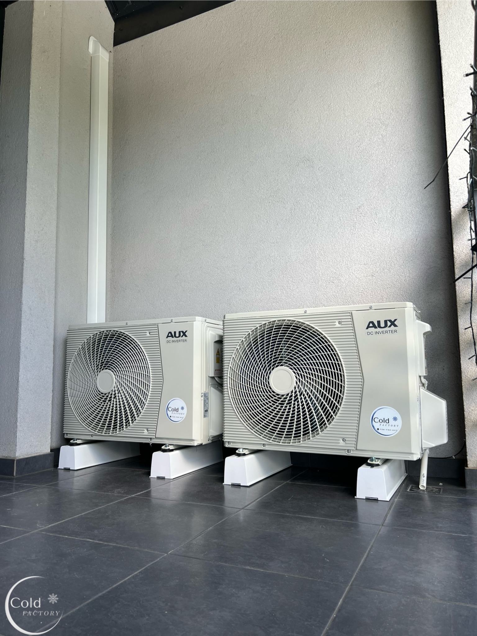 Dwa klimatyzatory AUX DC Inverter zamontowane na białych wspornikach na ciemnej, kafelkowanej podłodze przy szarej ścianie zewnętrznej budynku.