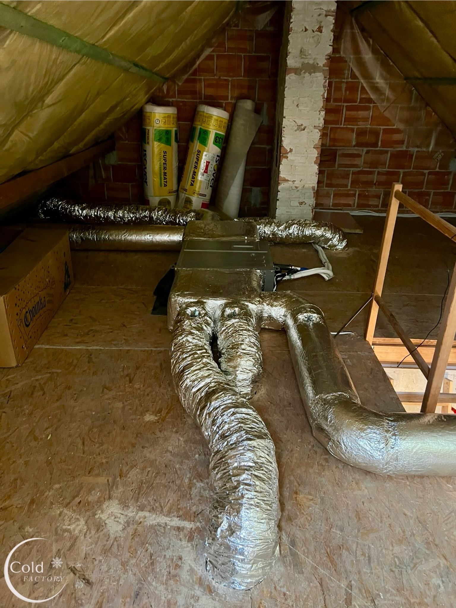 Instalacja rekuperacji na poddaszu: srebrne, izolowane rury wentylacyjne prowadzą do centrali rekuperacyjnej, widoczne materiały izolacyjne Isover, ceglany komin i kartonowe pudło Chojniak.