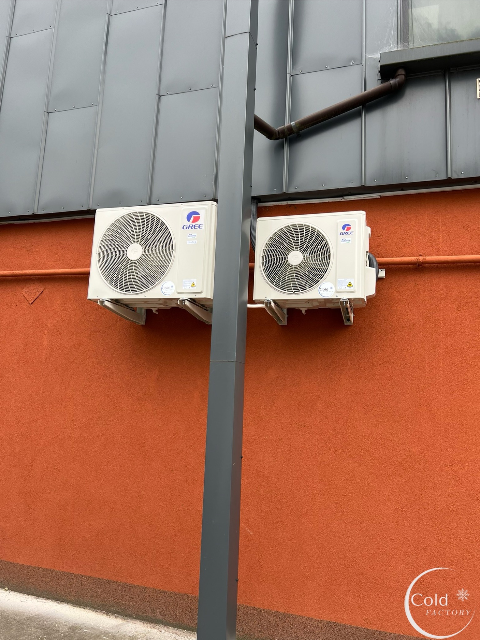 Dwa klimatyzatory marki Gree zamontowane na pomarańczowej ścianie budynku, widoczne rury spustowe i szara kolumna konstrukcyjna.
