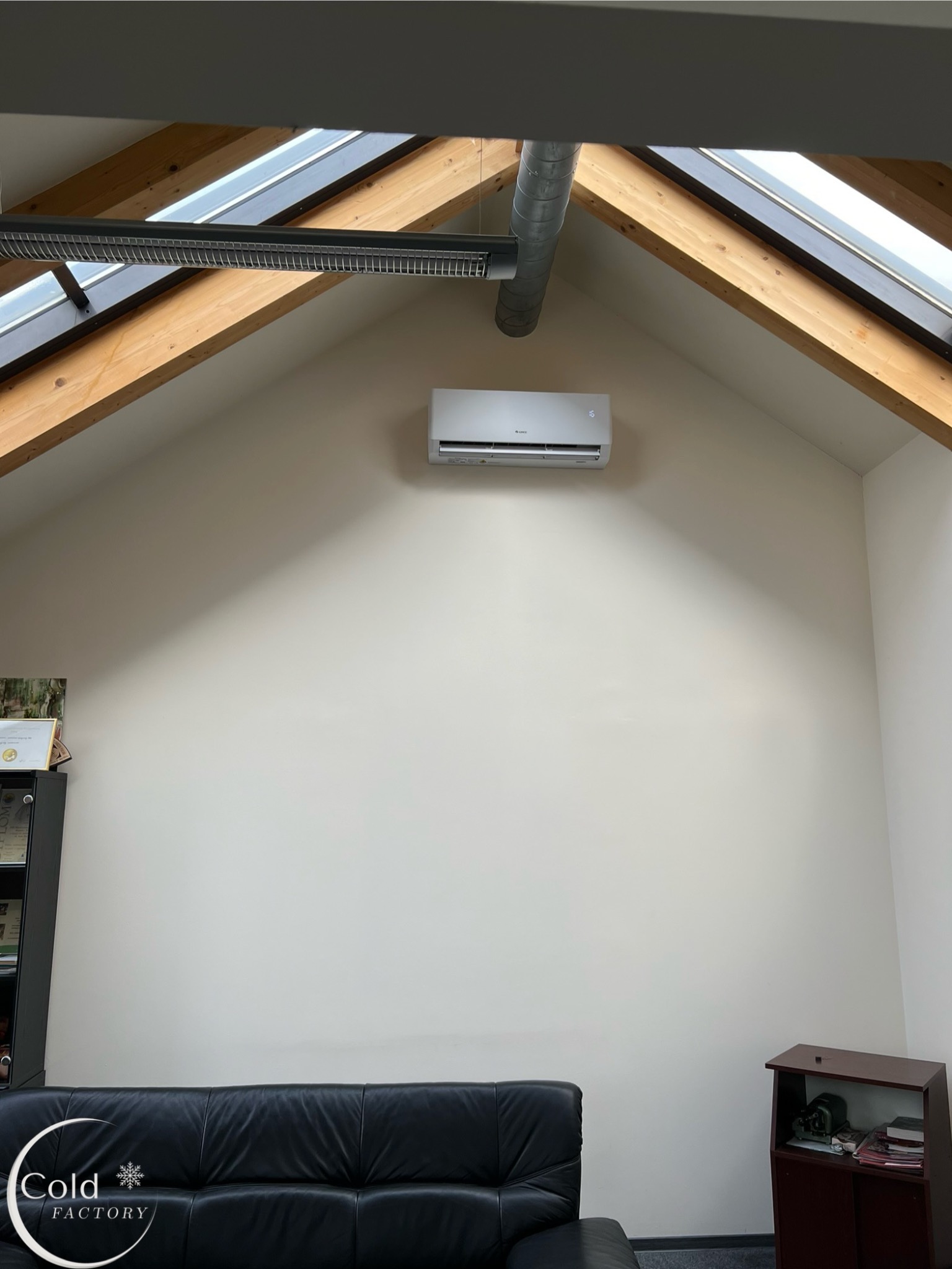 Wnętrze poddasza z zamontowaną białą jednostką klimatyzacji Gree na ścianie, widoczne drewniane belki stropowe, kratka wentylacyjna i fragment czarnej skórzanej sofy w dolnej części kadru.