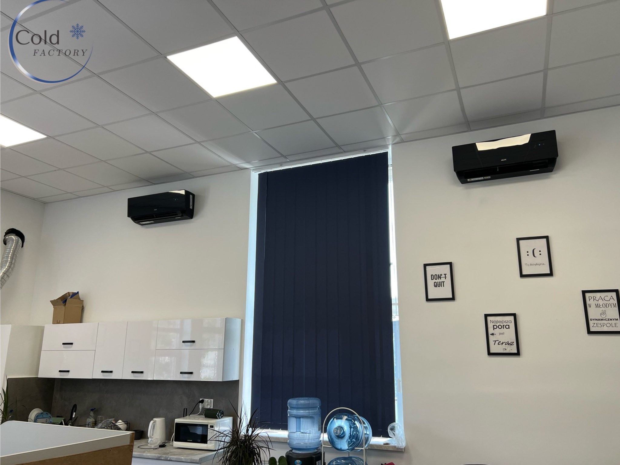 Dwa czarne klimatyzatory ścienne marki AUX w biurze z białymi ścianami, roletami w oknie, galerią motywacyjnych obrazków i aneksem kuchennym z białymi szafkami.