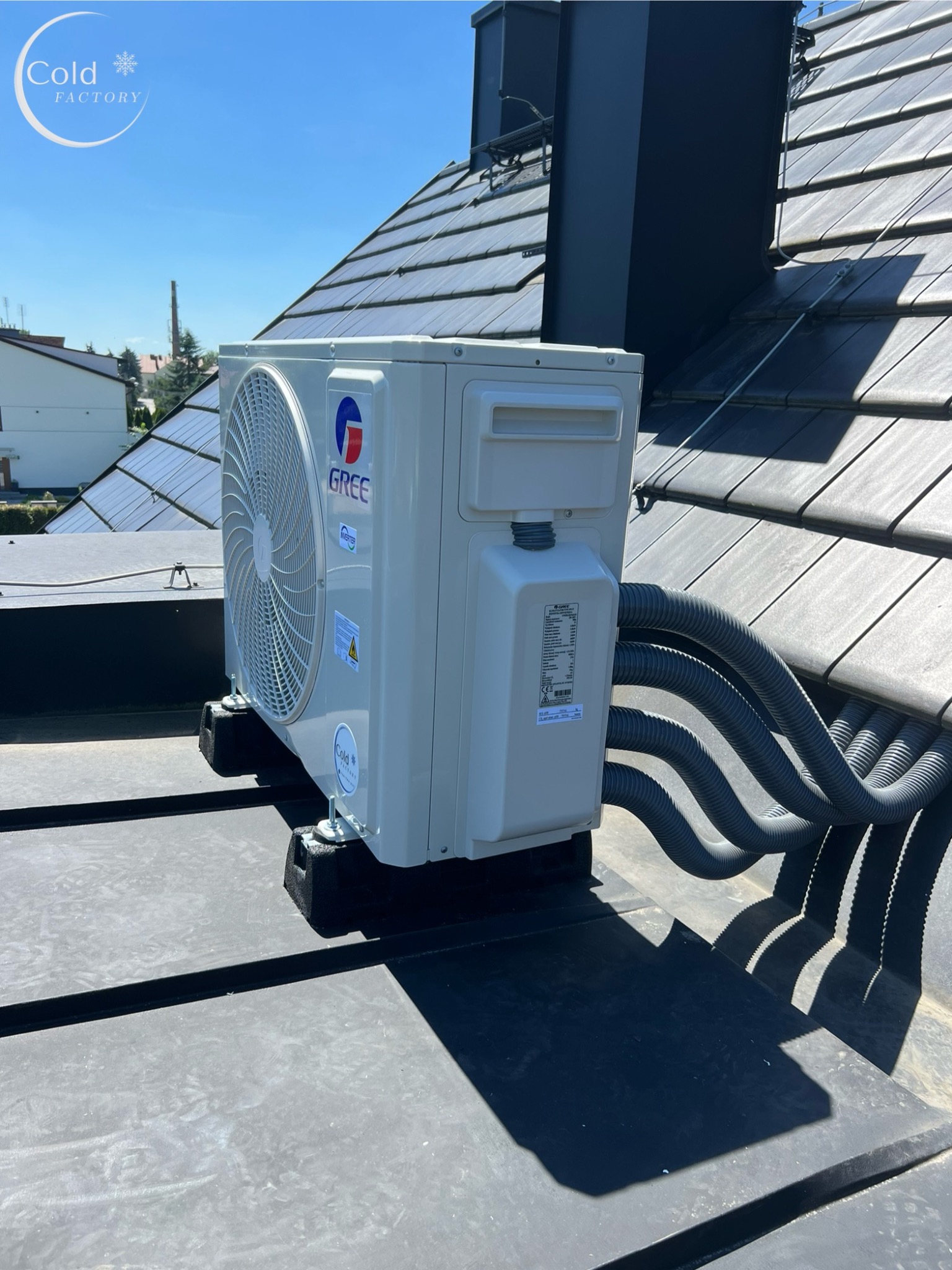 Zewnętrzna jednostka klimatyzacji marki Gree zamontowana na płaskim dachu, z widocznymi przewodami w szarych peszlach i elementami mocującymi.