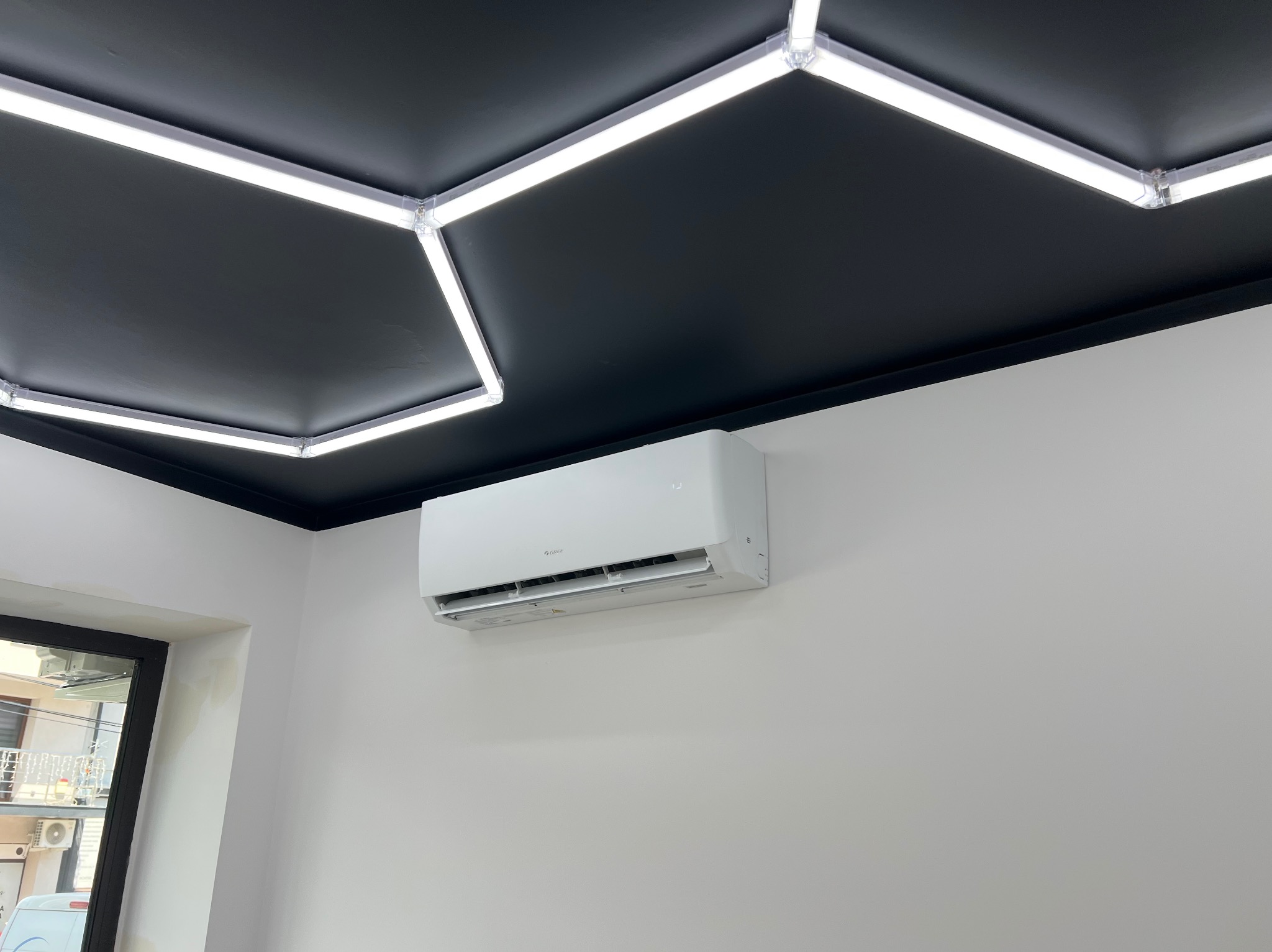 Nowoczesny klimatyzator Gree na białej ścianie, pod czarnym sufitem z geometrycznym oświetleniem LED. Widok z dołu, z fragmentem okna po lewej.