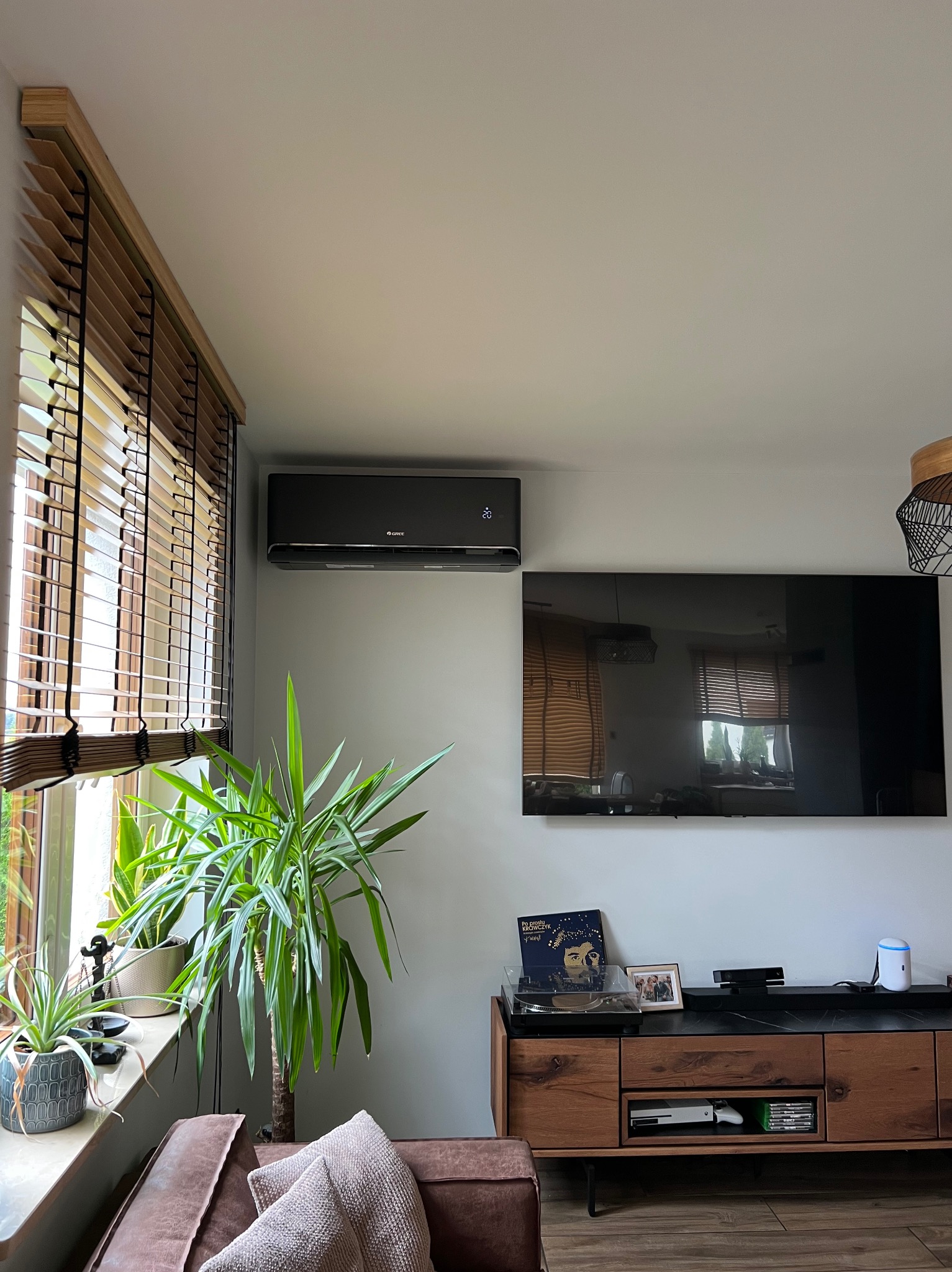 Nowoczesny, czarny klimatyzator Gree zamontowany na ścianie w salonie. Widoczne rolety drewniane, duży telewizor i rośliny doniczkowe. Minimalistyczny design.