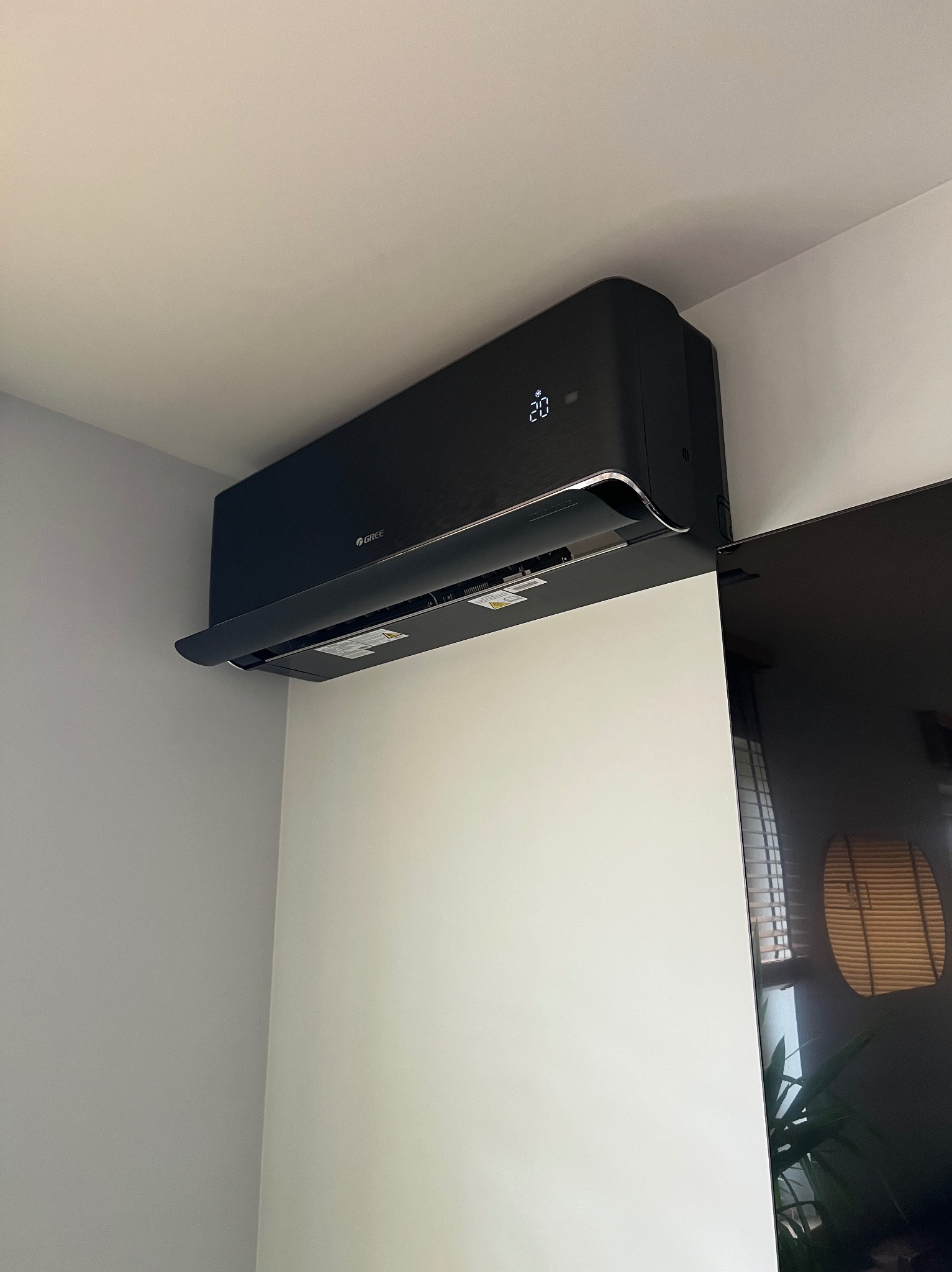 Czarna klimatyzacja Gree na ścianie, wyświetlająca temperaturę 20 stopni. Widoczny fragment telewizora i odbicie okna. Minimalistyczny design.