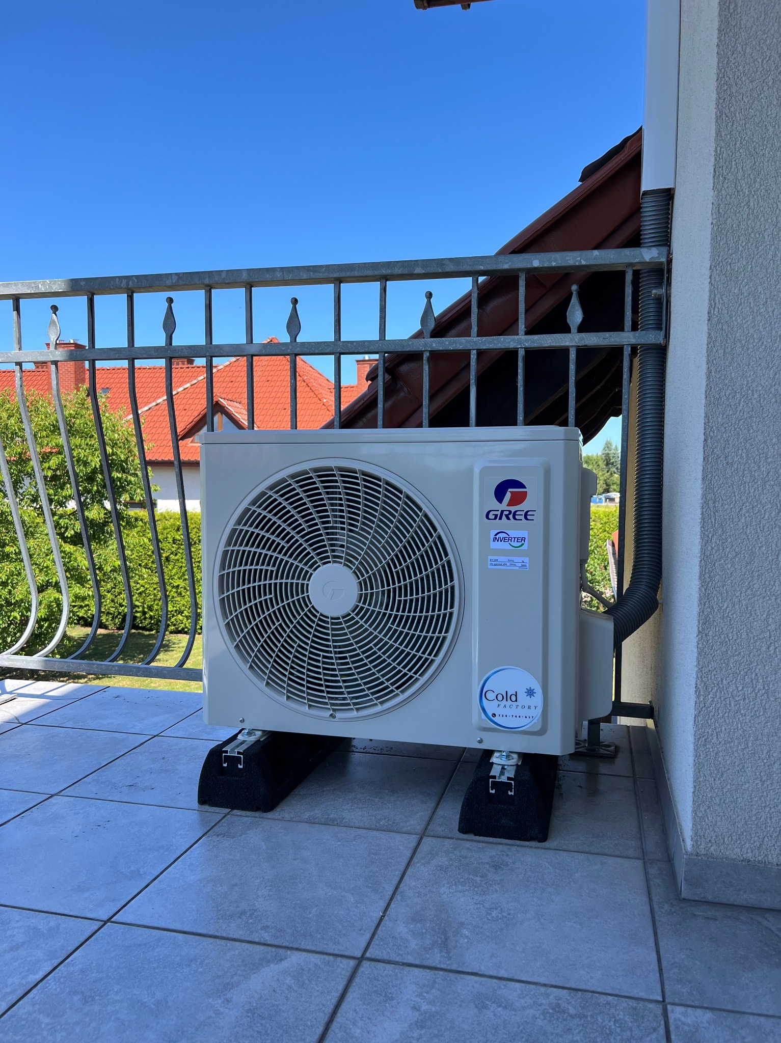 Klimatyzator Gree Inverter zamontowany na balkonie z szarą balustradą, na tle błękitnego nieba i czerwonego dachu sąsiedniego budynku. Widoczne elementy instalacji.