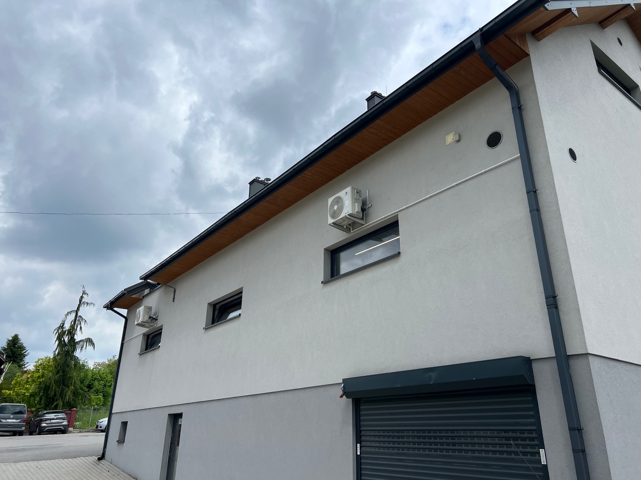 Szara elewacja budynku z zamontowanymi klimatyzatorami, widoczne okna z czarnymi ramami, brama garażowa, rynna i drewniane wykończenie dachu. Pochmurne niebo w tle.