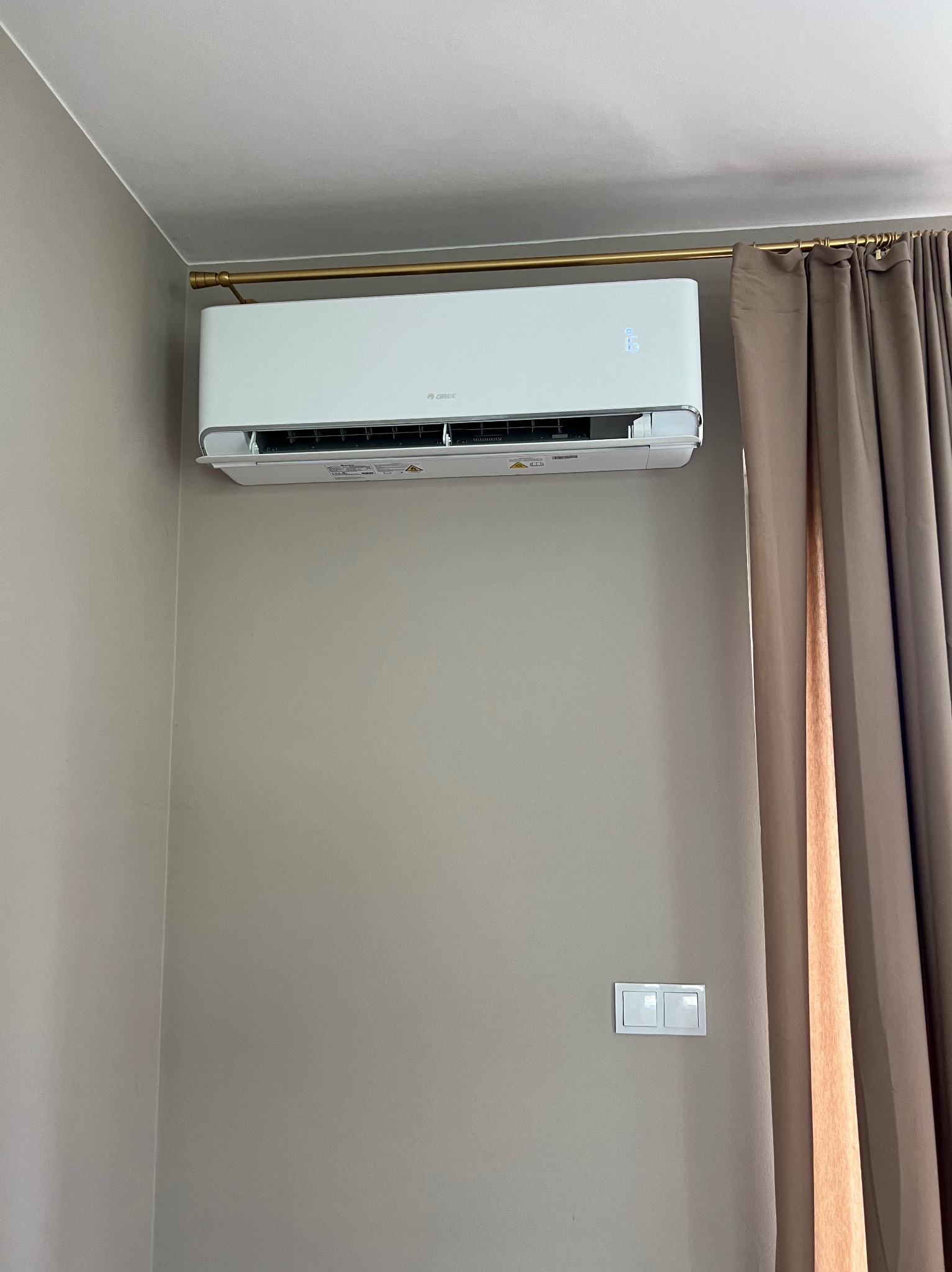 Biały klimatyzator Gree zamontowany na ścianie w pokoju, widoczne brązowe zasłony i włącznik światła. Minimalistyczny design wnętrza.