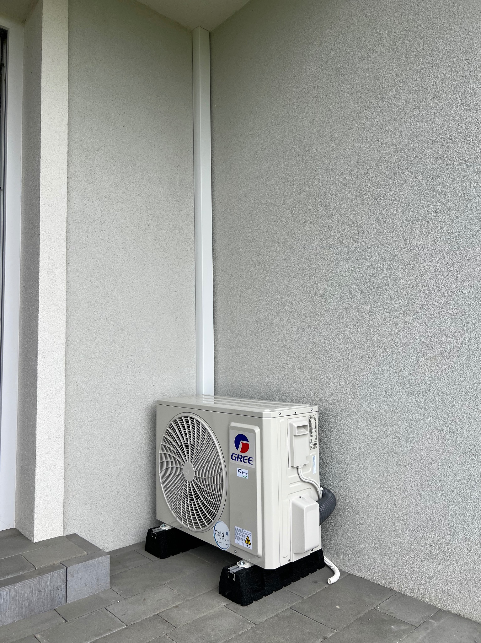 Zewnętrzna jednostka klimatyzacji Gree na czarnych podstawkach, umieszczona na szarym bruku przy ścianie z elewacją w jasnym odcieniu szarości. Widoczne elementy instalacji.