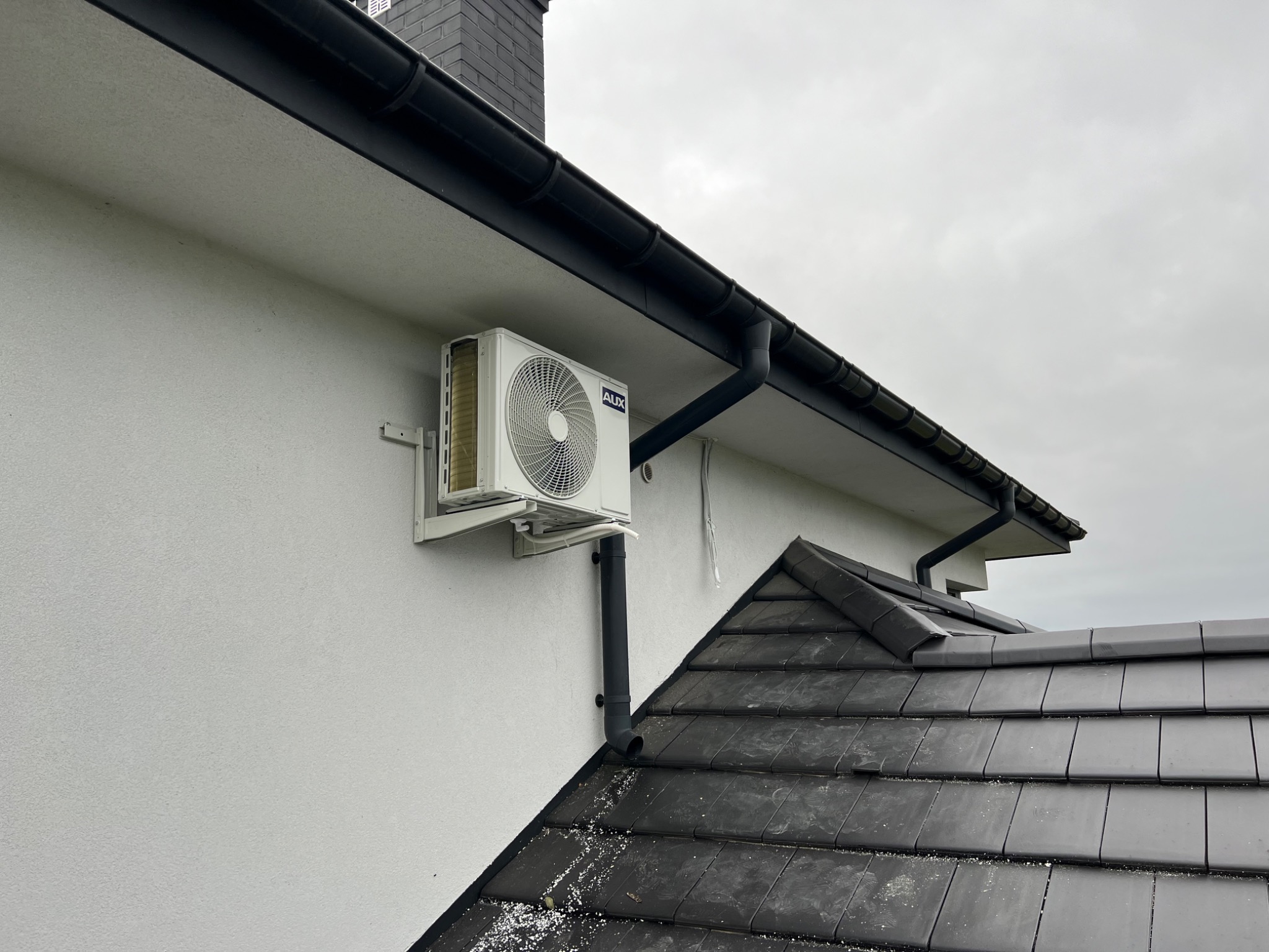Biała jednostka zewnętrzna klimatyzacji marki AUX zamontowana na elewacji domu pod czarną rynną, widok z dołu na tle szarego nieba.