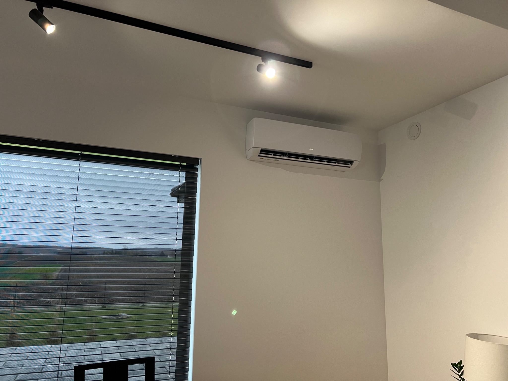 Biała jednostka klimatyzacyjna zamontowana na ścianie w jasnym pomieszczeniu z widokiem na krajobraz przez okno z żaluzjami.