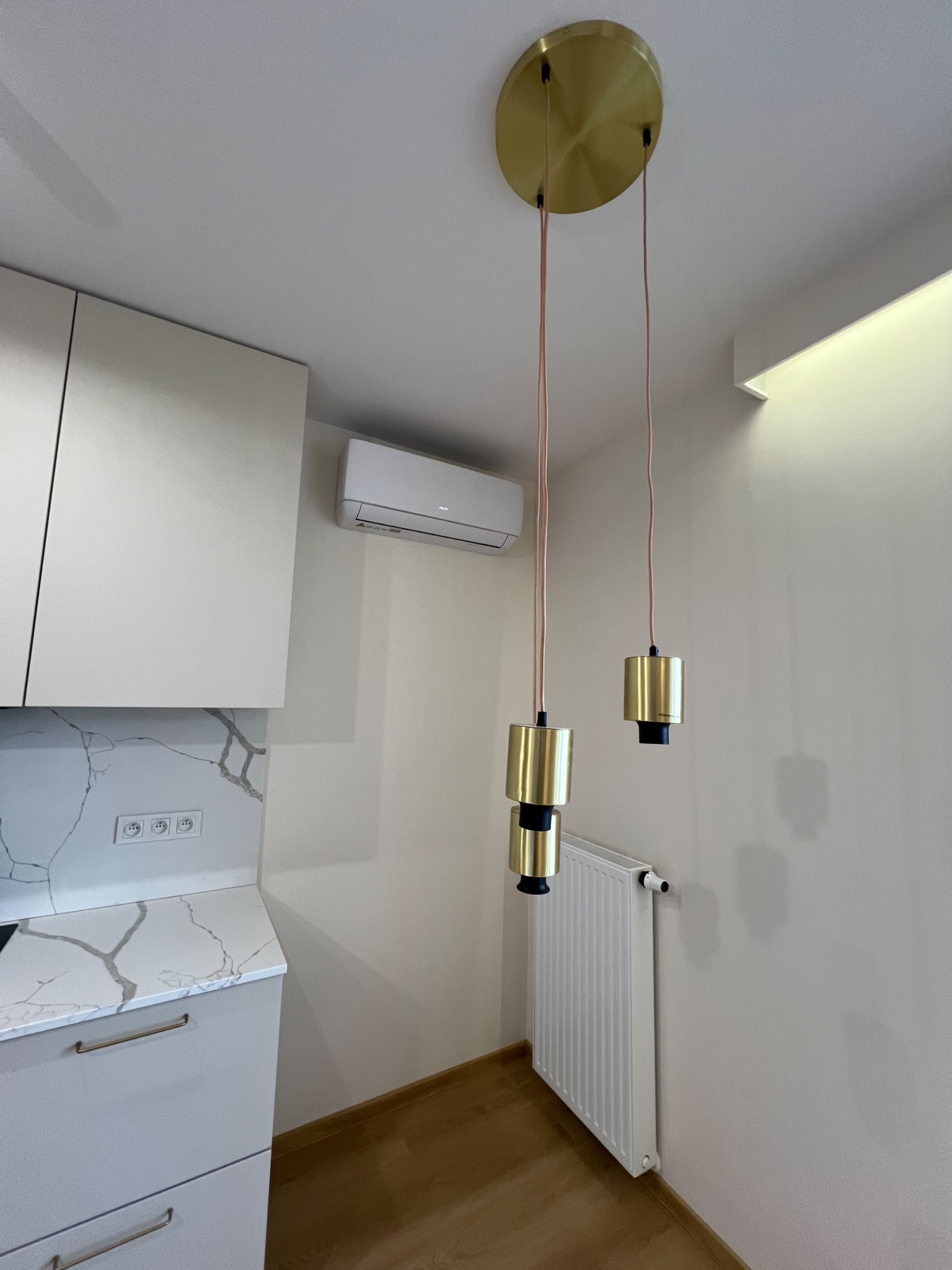 Klimatyzator ścienny zamontowany w kuchni nad blatem z imitacją marmuru, obok grzejnika i lamp wiszących z elementami w kolorze złotym.