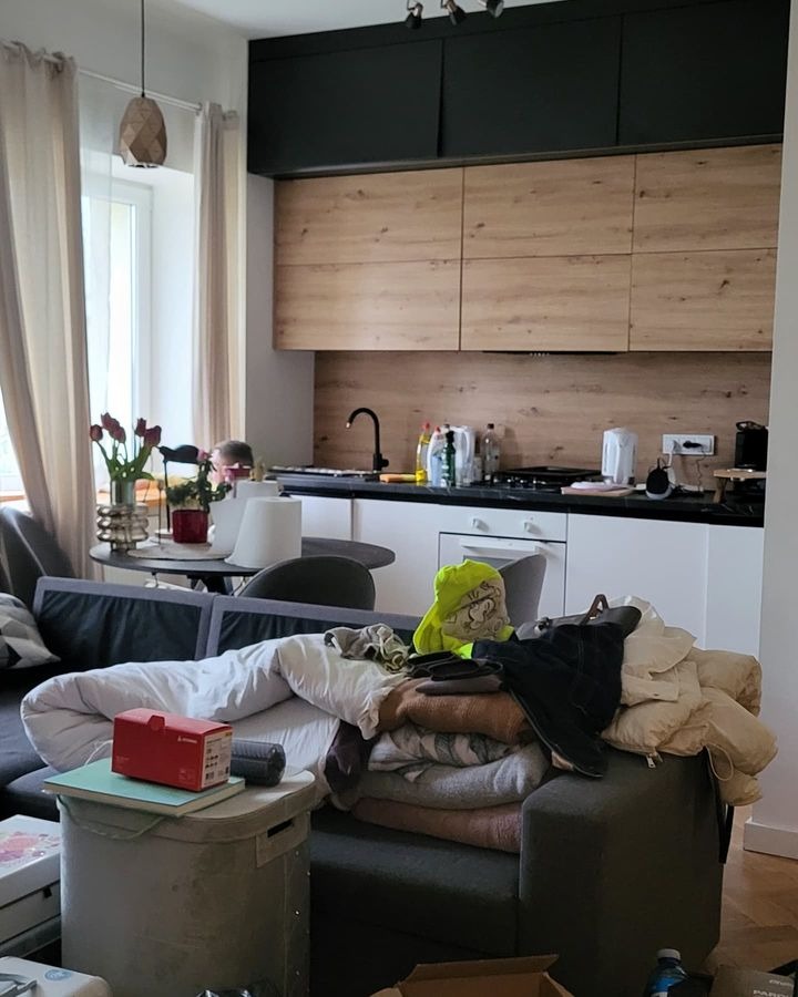 Zabałaganione mieszkanie w Lublinie: sterta ubrań i pościeli na kanapie na tle kuchni z drewnianymi szafkami.