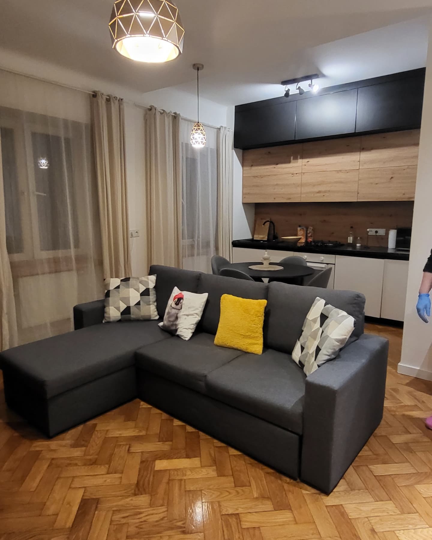 Szary narożnik z poduszkami w salonie z parkietem w jodełkę, widoczna kuchnia w tle z drewnianymi frontami i czarnymi szafkami wiszącymi, oświetlenie sufitowe.