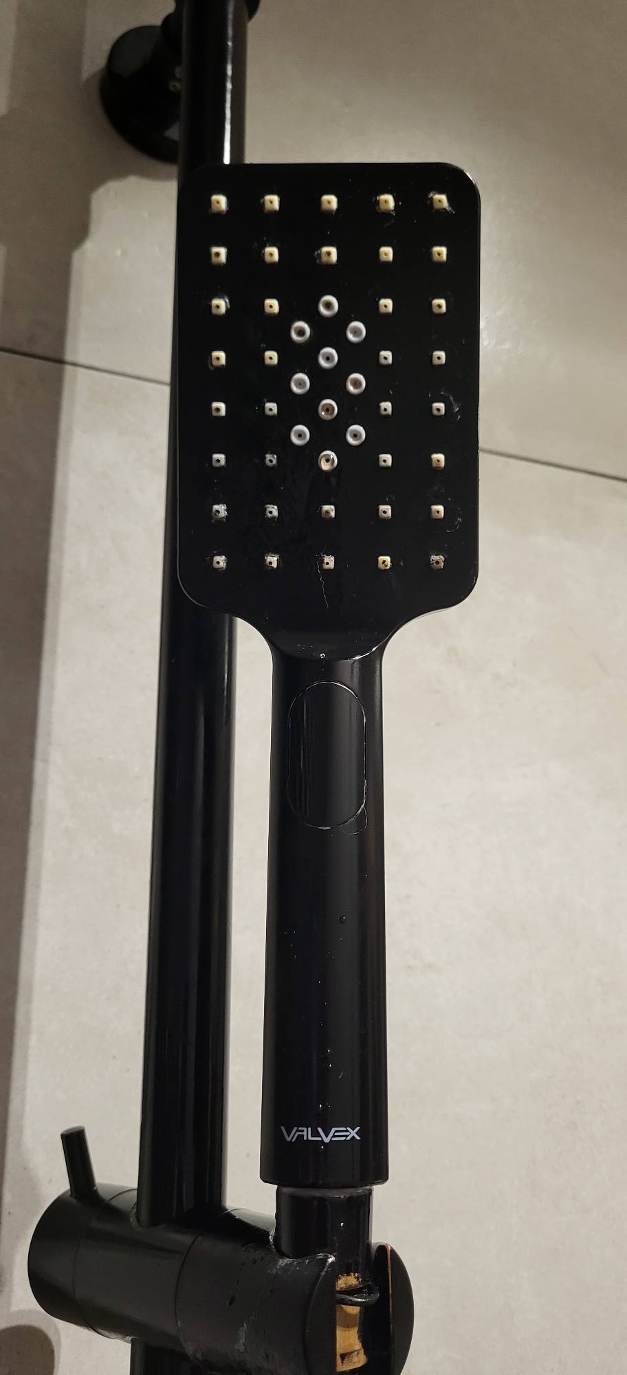 Czarna, kwadratowa deszczownica z widocznym logo Valvex, zamontowana na ścianie z jasnych płytek.