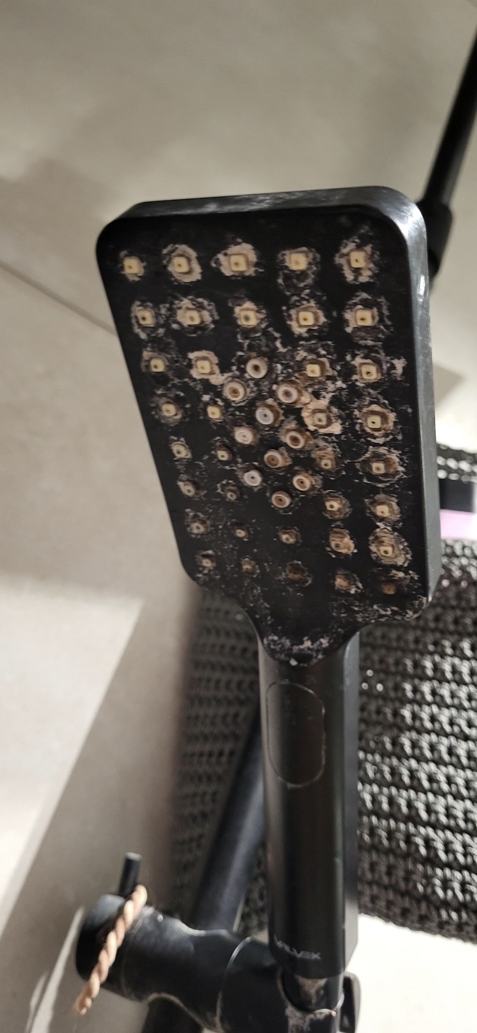 Zakamieniona, czarna słuchawka prysznicowa z dyszami zapchanymi osadem, widoczny wężyk prysznicowy i fragment ściany.