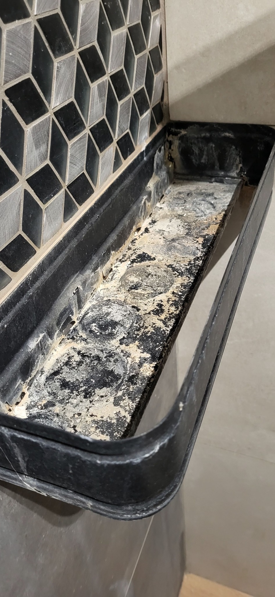 Silnie zabrudniona, czarna półka narożna w kabinie prysznicowej, z widocznymi osadami kamienia i resztkami mydła, na tle mozaiki z rombów.