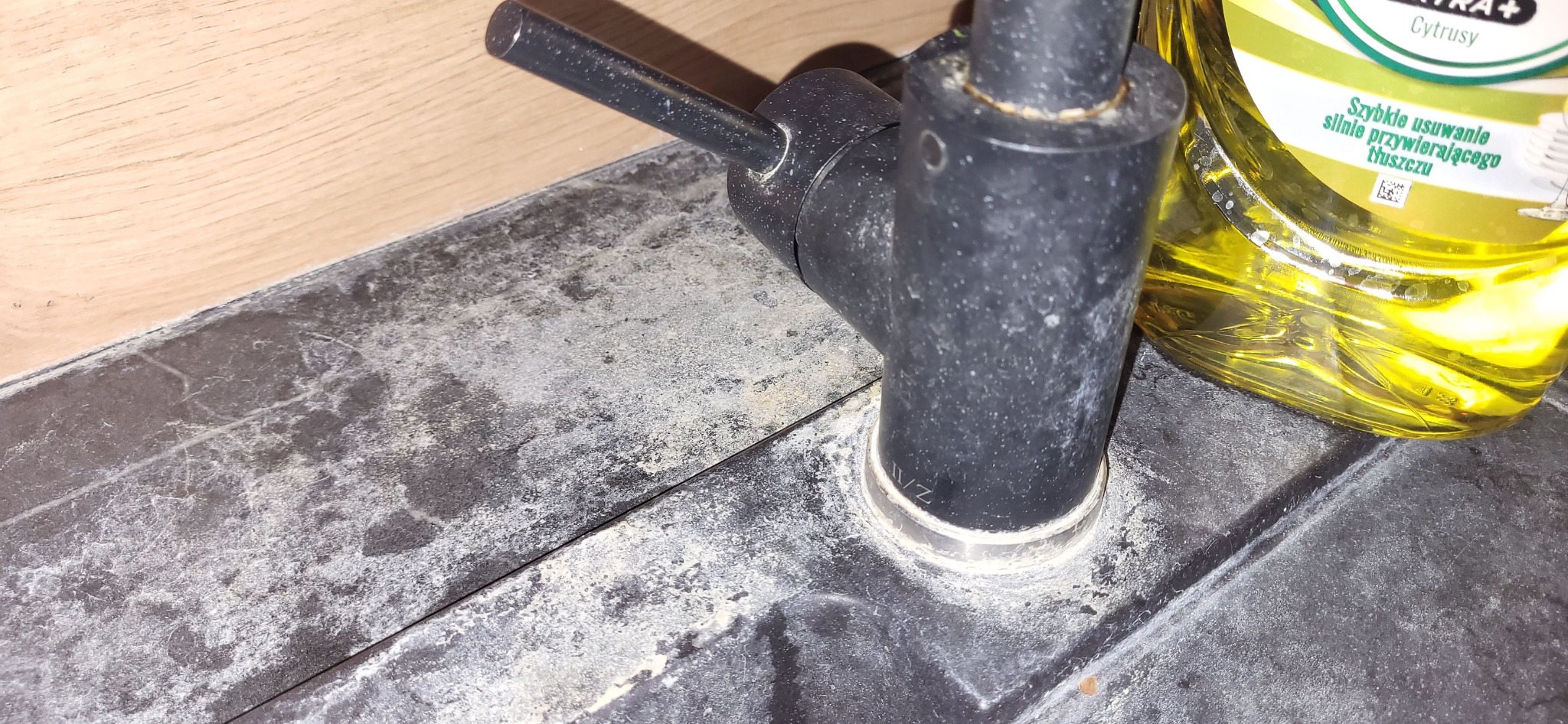 Czarny kran z osadem kamiennym na zlewie, obok butelka płynu do mycia naczyń z napisem 'Szybkie usuwanie silnie przywierającego tłuszczu'.