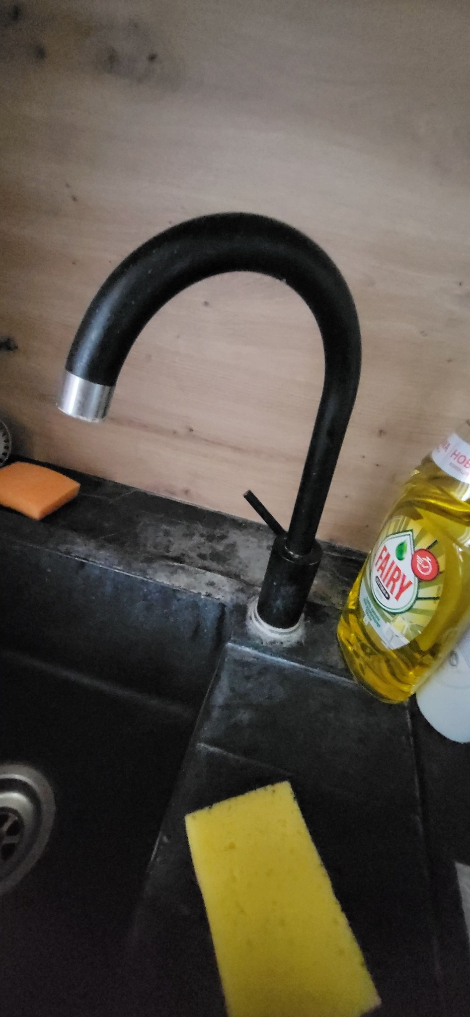 Czarny kran nad czarnym zlewem, obok gąbka i płyn do naczyń Fairy, widoczne ślady użytkowania na zlewie.