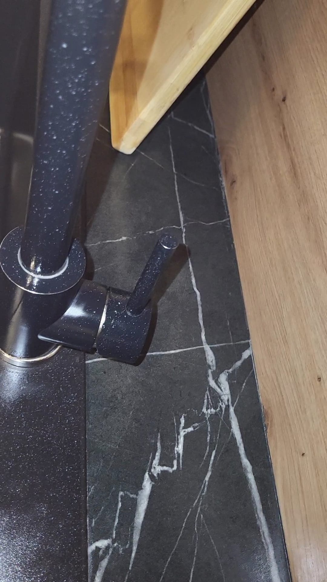 Czarny kran kuchenny z białymi plamkami na tle blatu z czarnego marmuru z białymi żyłami i fragmentu drewnianej półki.