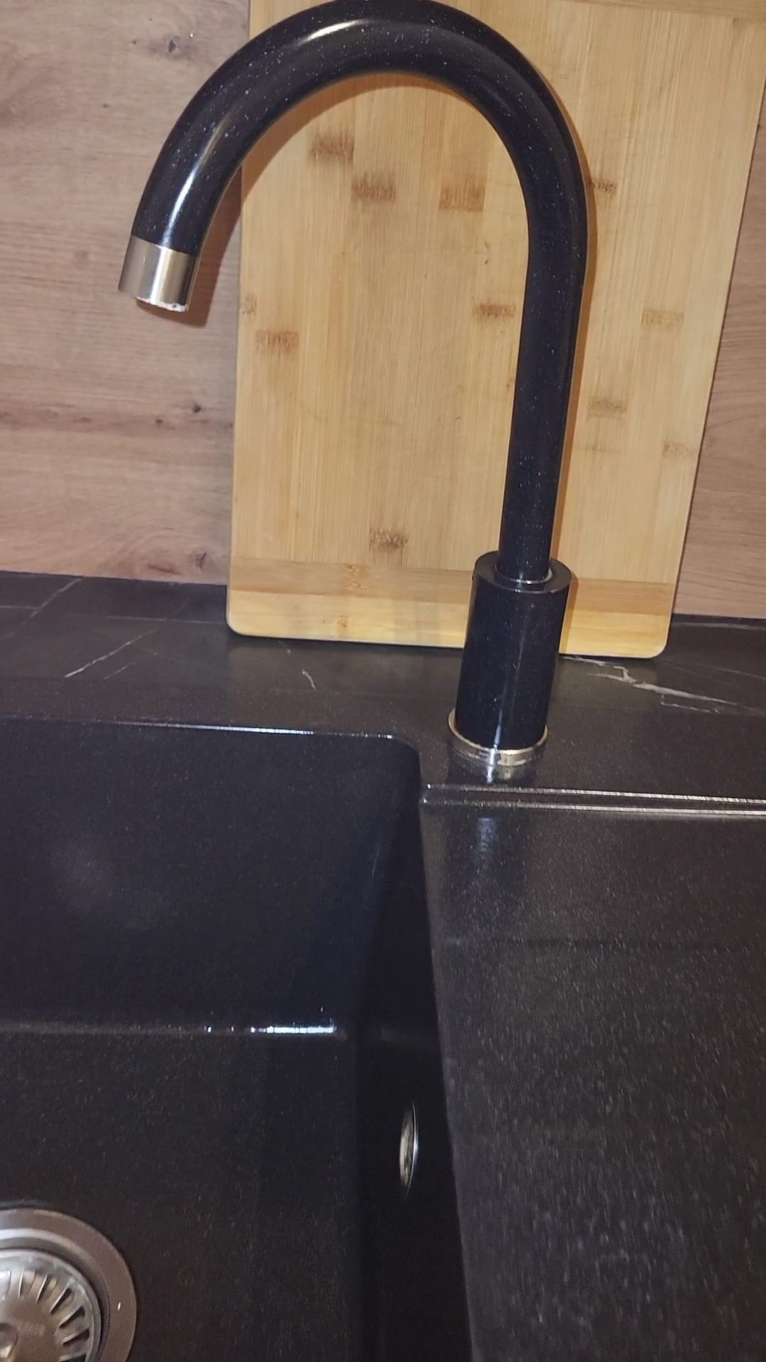 Czarny, granitowy zlew kuchenny z wysoką, zakrzywioną baterią z elementami chromowanymi, deska do krojenia w tle.