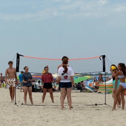 Biuro Turystyczne Skaut Tour sp. z o. o. - Młodzież gra w siatkówkę na piaszczystej plaży, w tle kolorowe parasole i tłum ludzi, siatka z pomarańczową górną krawędzią, pochmurne niebo.