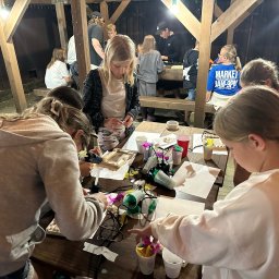 Biuro Turystyczne Skaut Tour sp. z o. o. - Warsztaty artystyczne dla dzieci: grupa dziewcząt tworzy ozdoby z piór i koralików przy drewnianym stole pod zadaszeniem, oświetlonym ciepłym światłem.