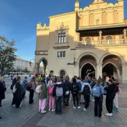 Wycieczka szkolna na Rynku Głównym w Krakowie. Grupa uczniów z plecakami stoi przed Sukiennicami w słoneczny dzień. Widoczna wieża kościoła Mariackiego.