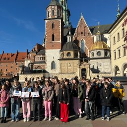 Biuro Turystyczne Skaut Tour sp. z o. o. - Wycieczka szkolna z Łodzi na tle Wawelu. Grupa uczniów trzymająca tabliczki z napisem 'Podróże z klasą' w słoneczny dzień.